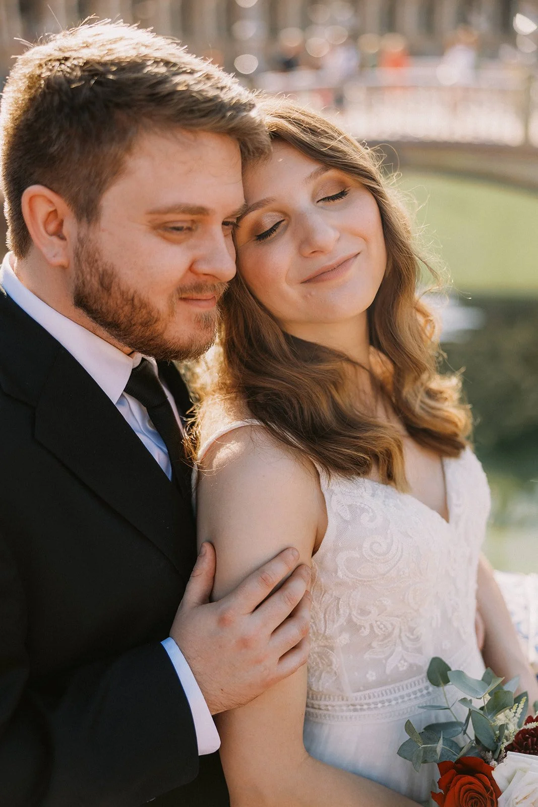 A couple on their wedding day, pose closely with their heads touching. The man wears a black tuxedo and the woman wears a white lace dress. They appear happy in a scenic outdoor setting with a bridge and water in the background.