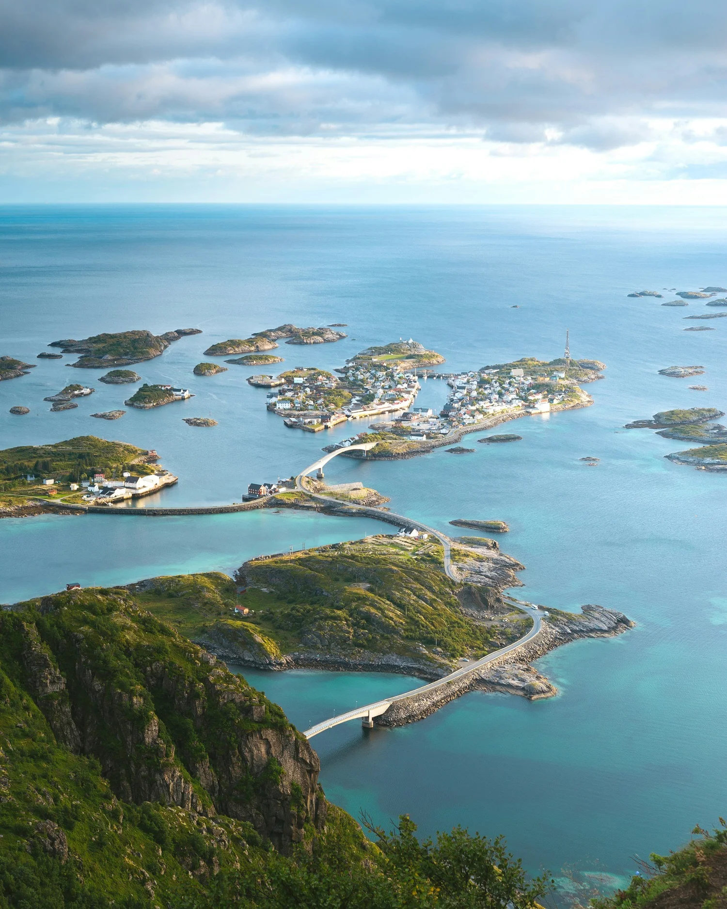 Aerial view of a coastal area with green cliffs, a town, and a winding bridge over the water, under a cloudy sky.