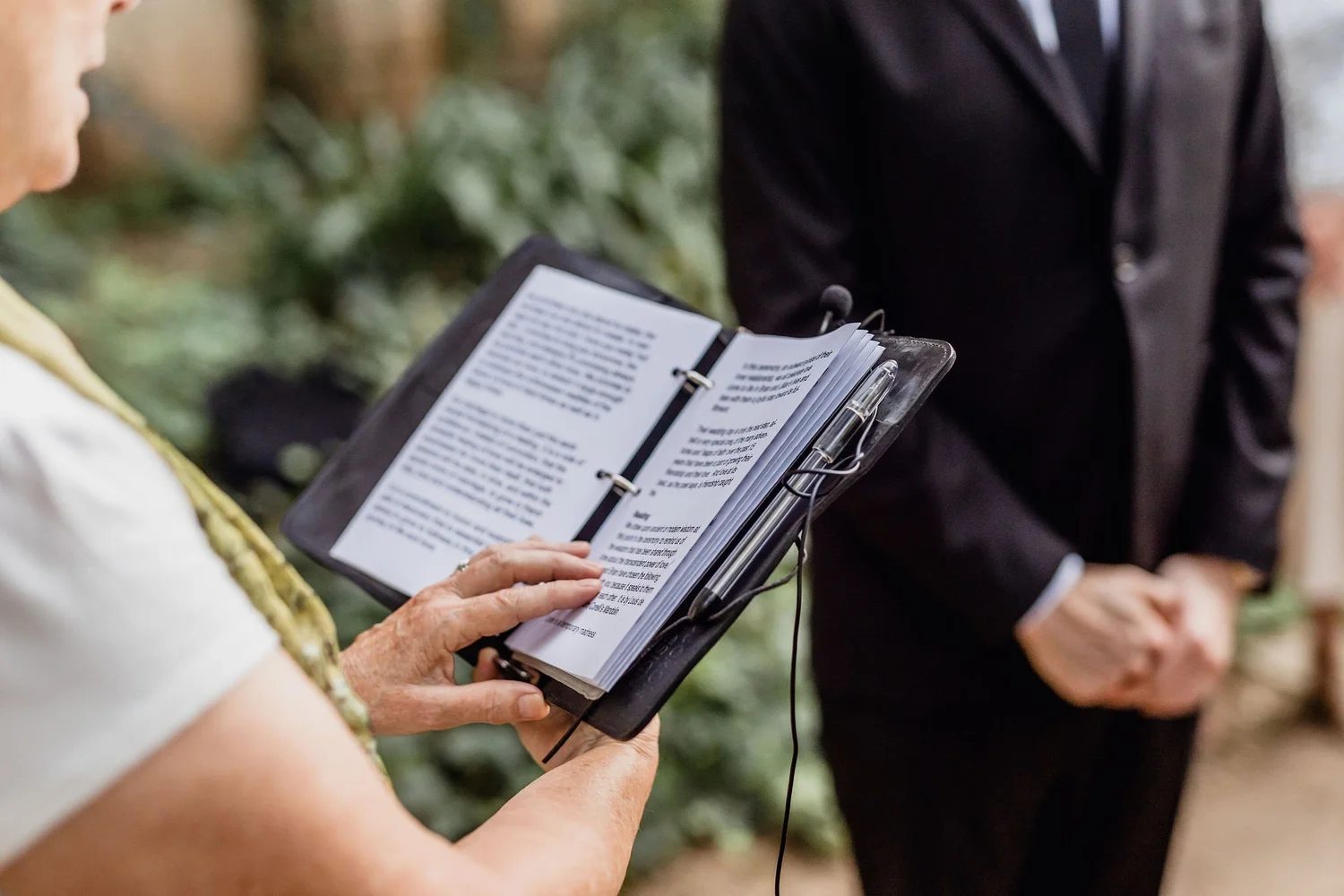 A person holding a booklet or pamphlet reading from it, while another person in a black suit looks on, in an outdoor setting with blurred greenery in the background.