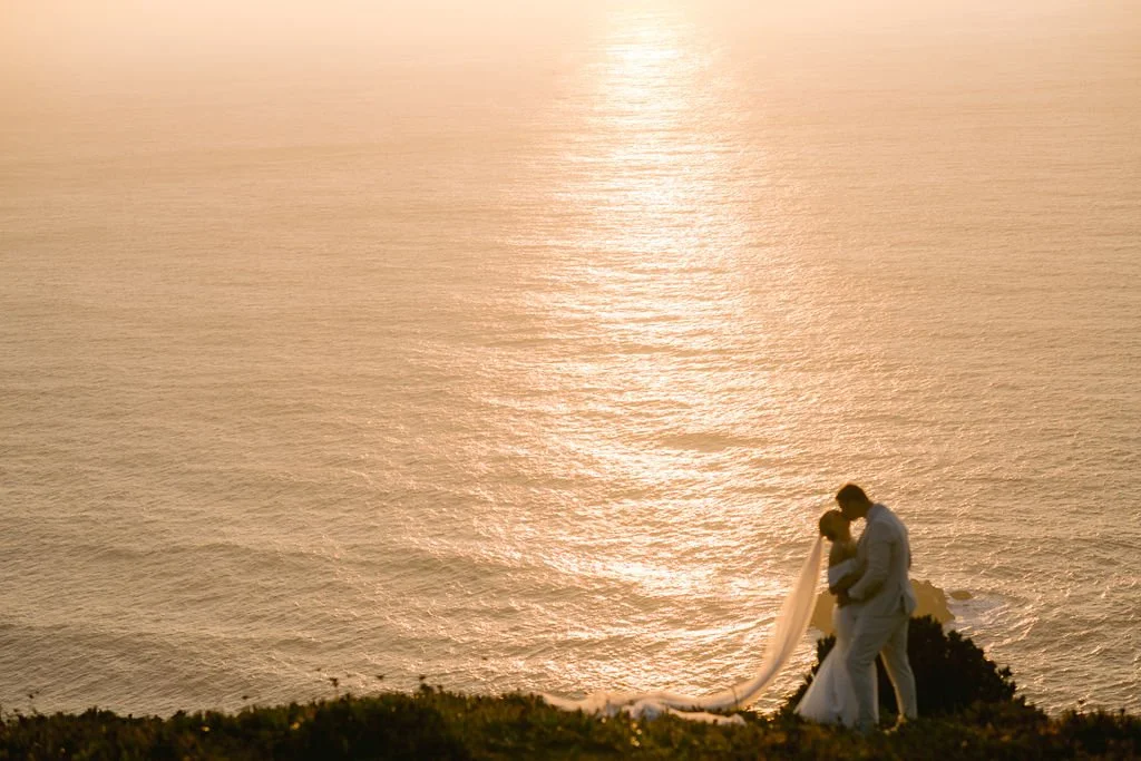 A couple gets married on a cliff overlooking the ocean during sunset, with the bride holding a flowing veil.