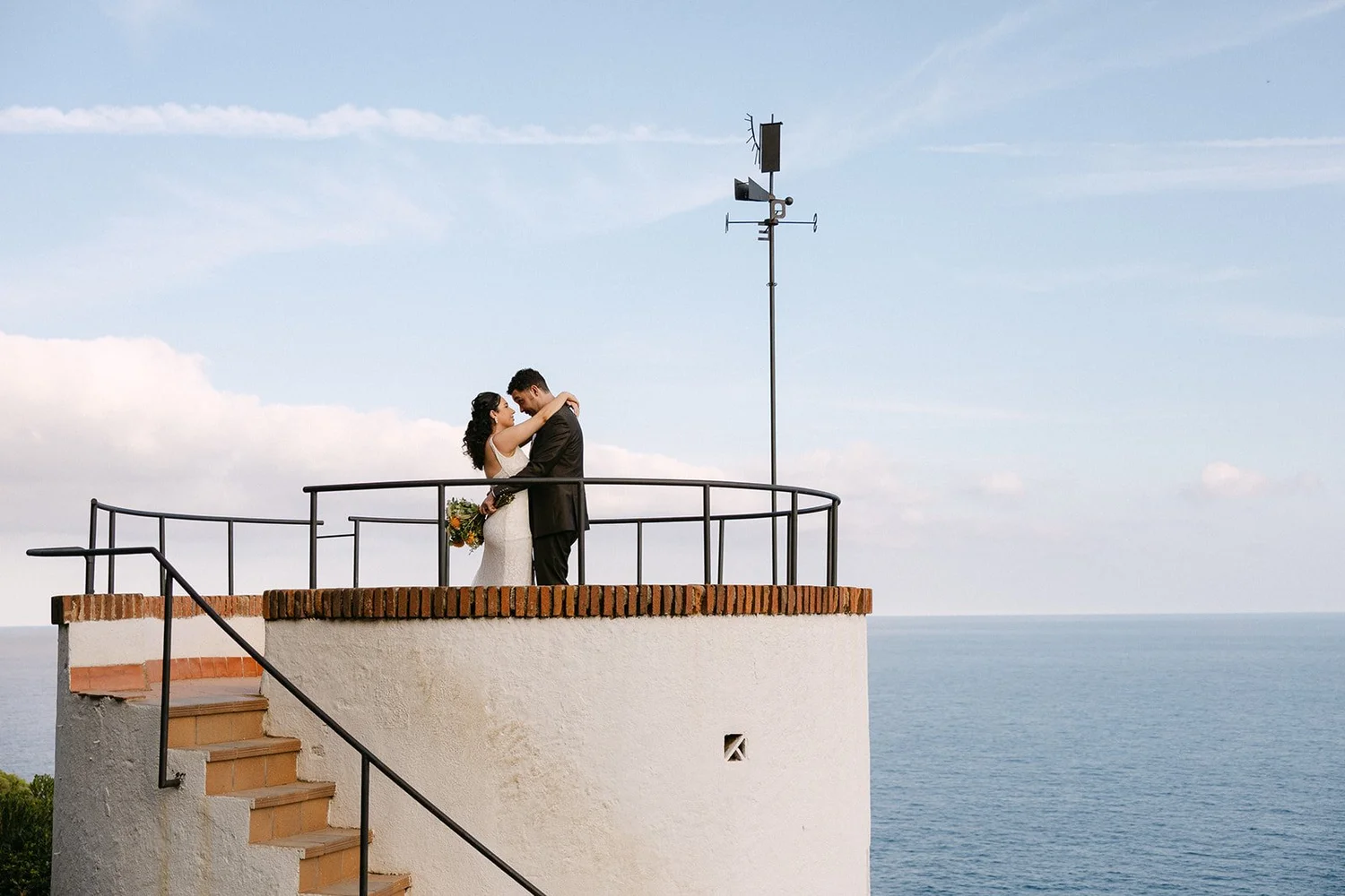 A couple in wedding attire embracing on a rooftop overlooking the ocean, with a blue sky and clouds in the background.