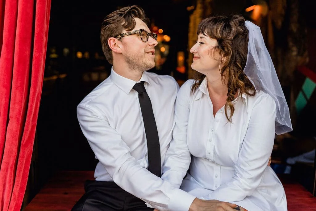 A couple, possibly newlyweds, sitting close together on a red bench, facing each other and smiling. The woman wears a white dress and a veil, and the man wears a white shirt, black tie, and glasses. They are in an intimate, joyful moment indoors with dim lighting and red curtains nearby.