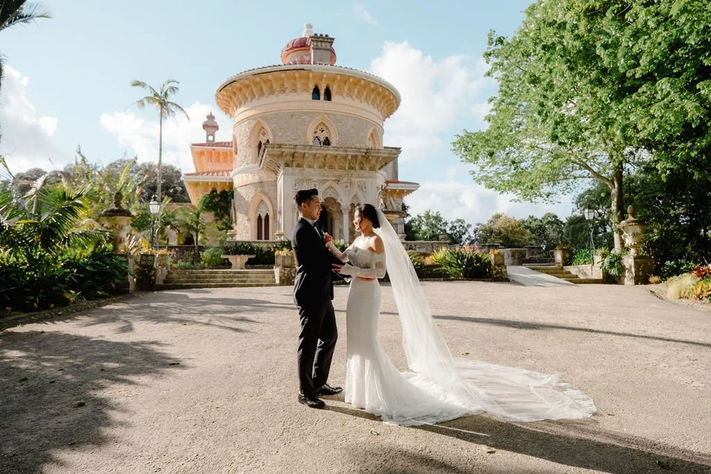 A bride and groom in wedding attire holding hands outside a grand, ornate building under a partly cloudy sky. The bride has a long veil and white gown, and the groom is in a tuxedo. There are trees and lush greenery around.