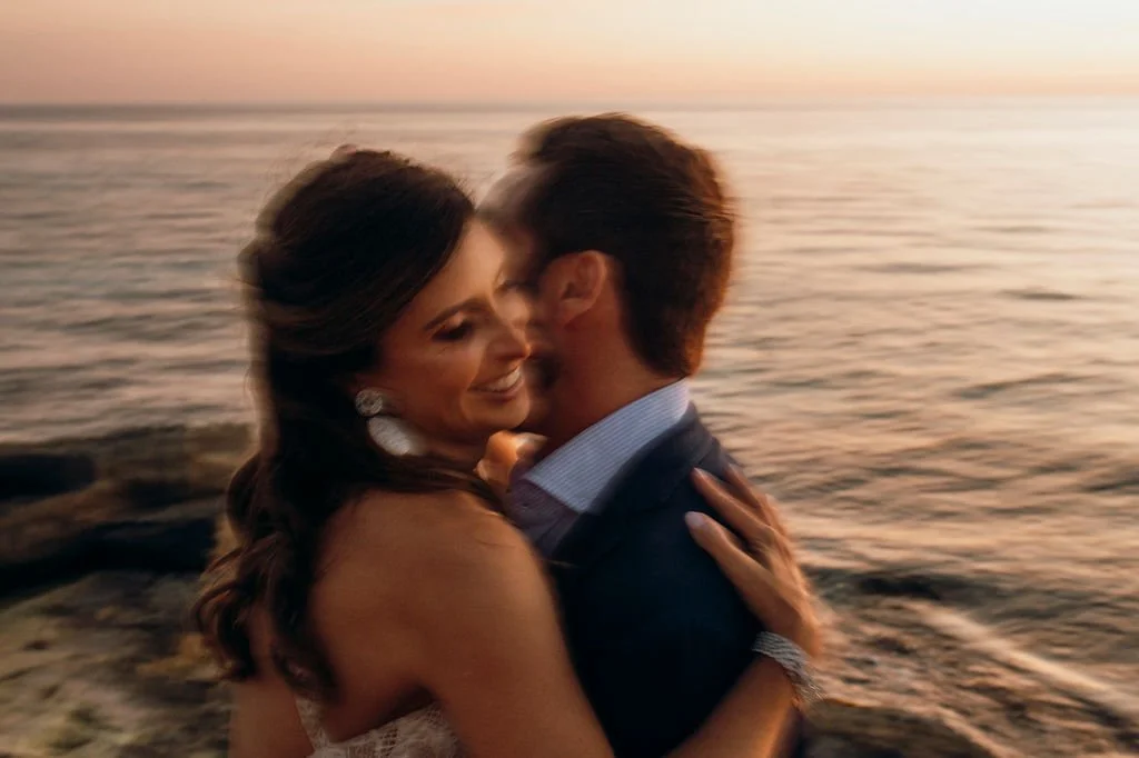 A smiling woman with earrings and long hair is embracing a man on a beach during sunset, both with eyes closed, near the water.