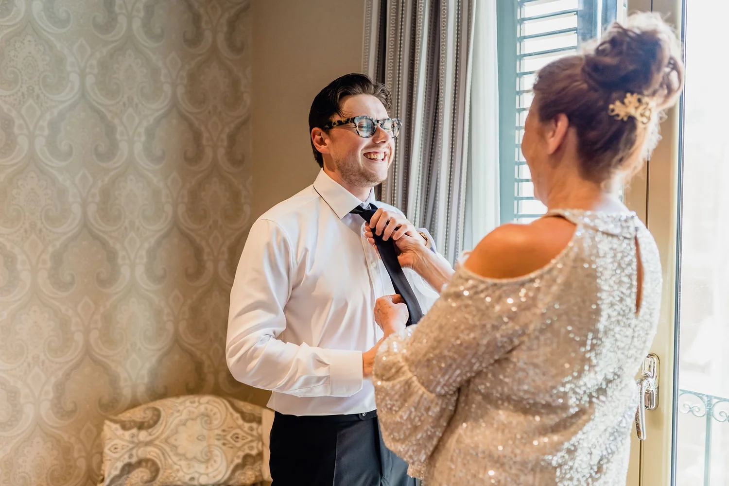 A young man with stylish glasses smiling as he adjusts his black tie, facing an older woman in an elegant, sparkling dress, in a well-lit room with patterned wallpaper and a window with blinds.