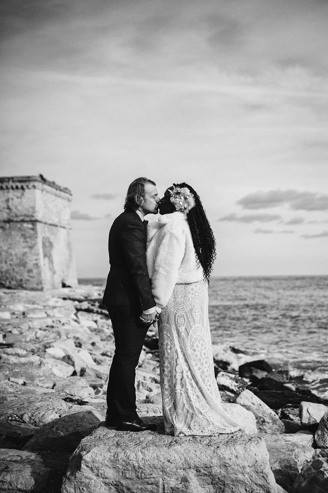 A black and white photo of a couple kissing on a rocky beach, holding hands, with a historic stone structure in the background.