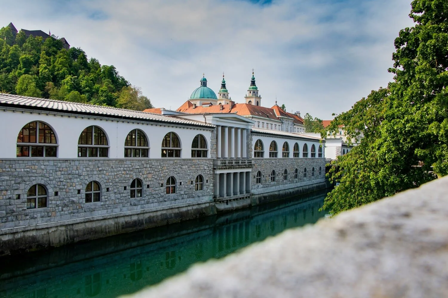 View of a historic building with arched windows and a stone facade beside a canal, with a church with green domes and spires in the background, surrounded by trees and a hillside.
