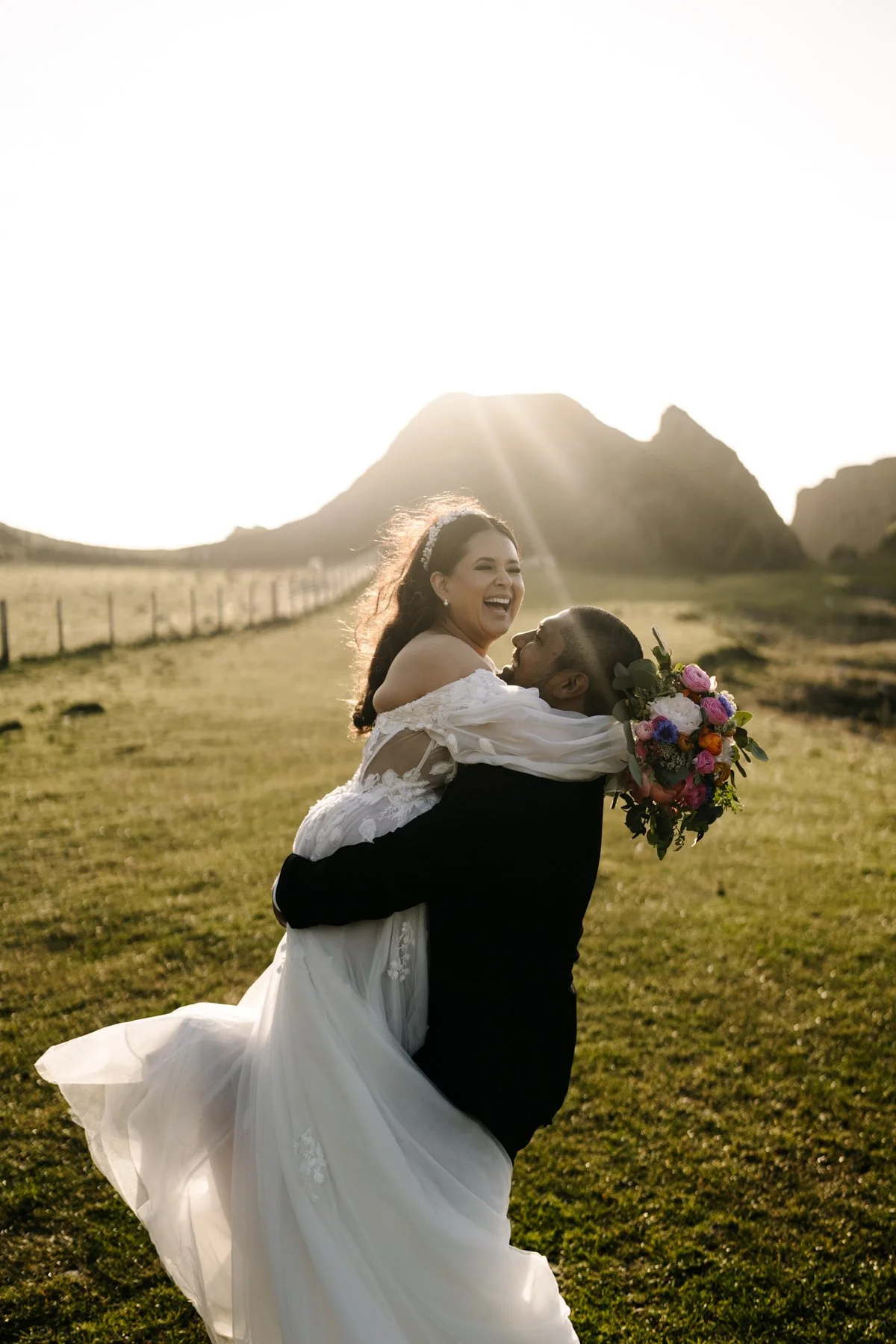 A couple on their wedding day, with the groom lifting the bride who is holding a bouquet of colorful flowers, in a grassy outdoor area with mountains in the background and the sun setting.