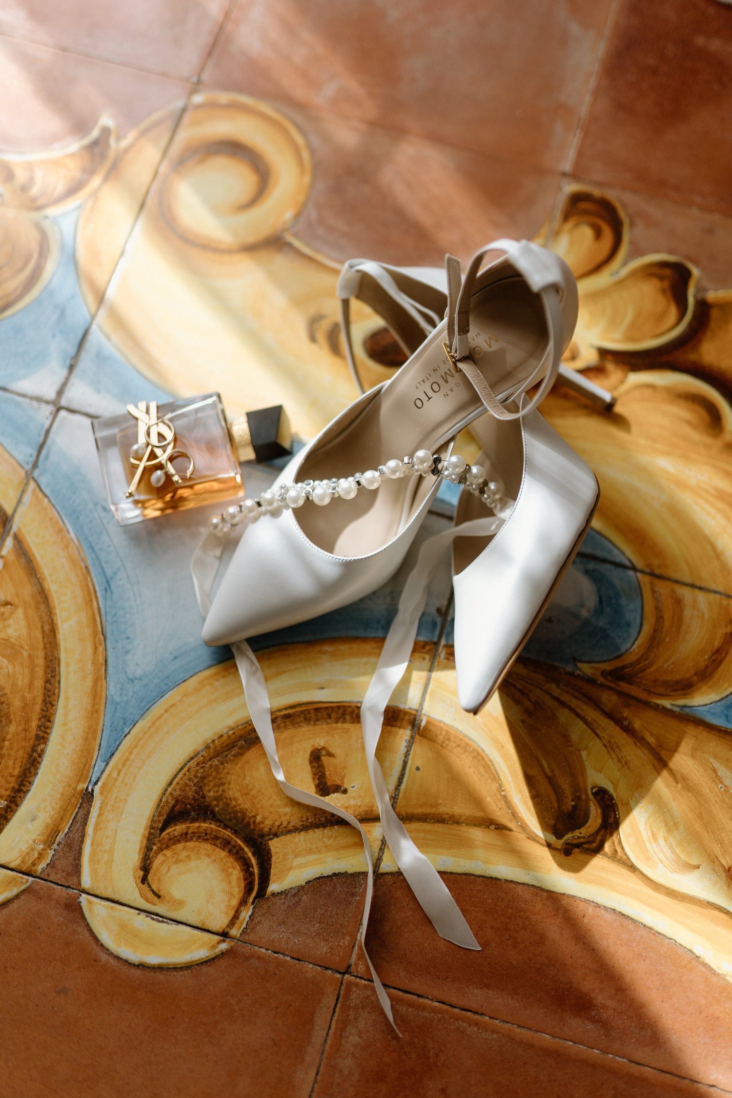 A pair of white high-heeled shoes with ankle straps, pearl necklace, a YSL perfume bottle, and a ring, all placed on a colorful tiled floor.