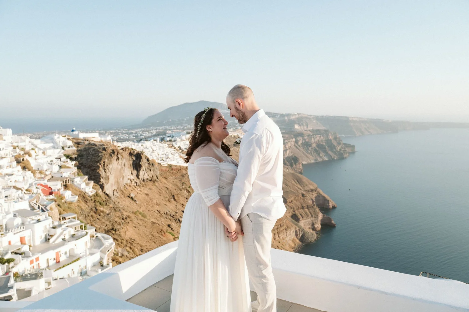 A couple dressed in wedding attire standing close together on a balcony overlooking white buildings, cliffs, and the sea in Santorini, Greece.