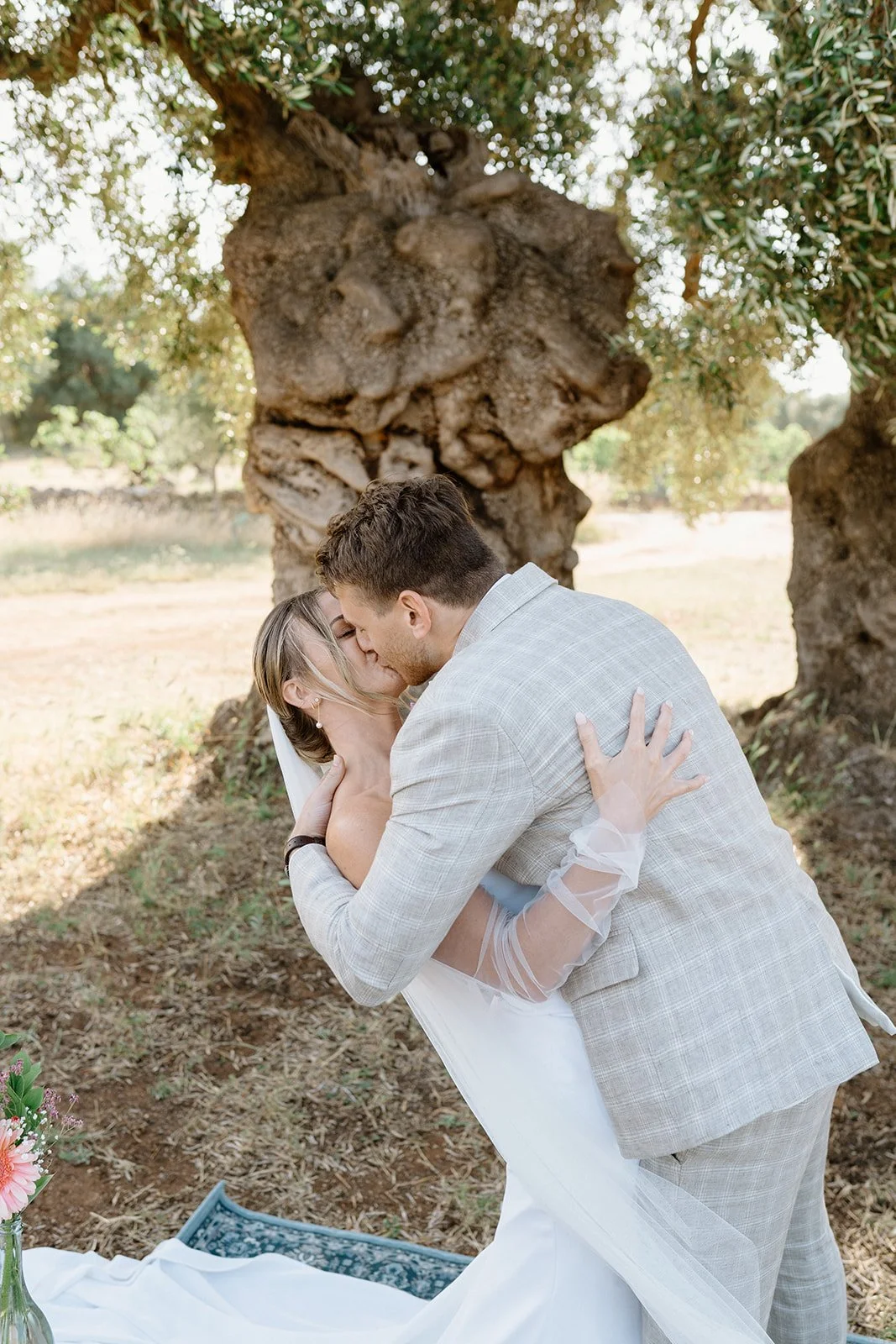 A couple in wedding attire sharing a kiss outdoors under a large tree during their wedding ceremony.