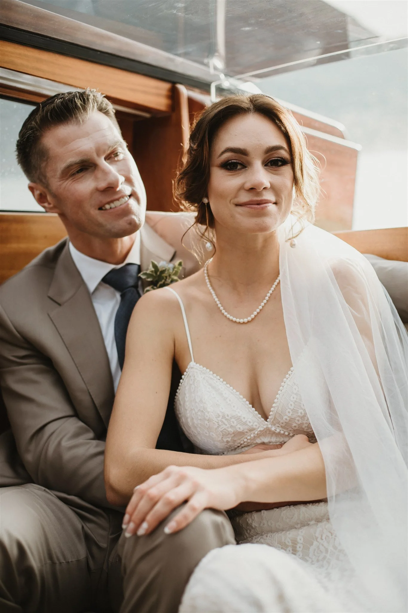 A happy couple dressed in wedding attire sitting closely together on a wooden bench, smiling at the camera in a warmly lit indoor setting.