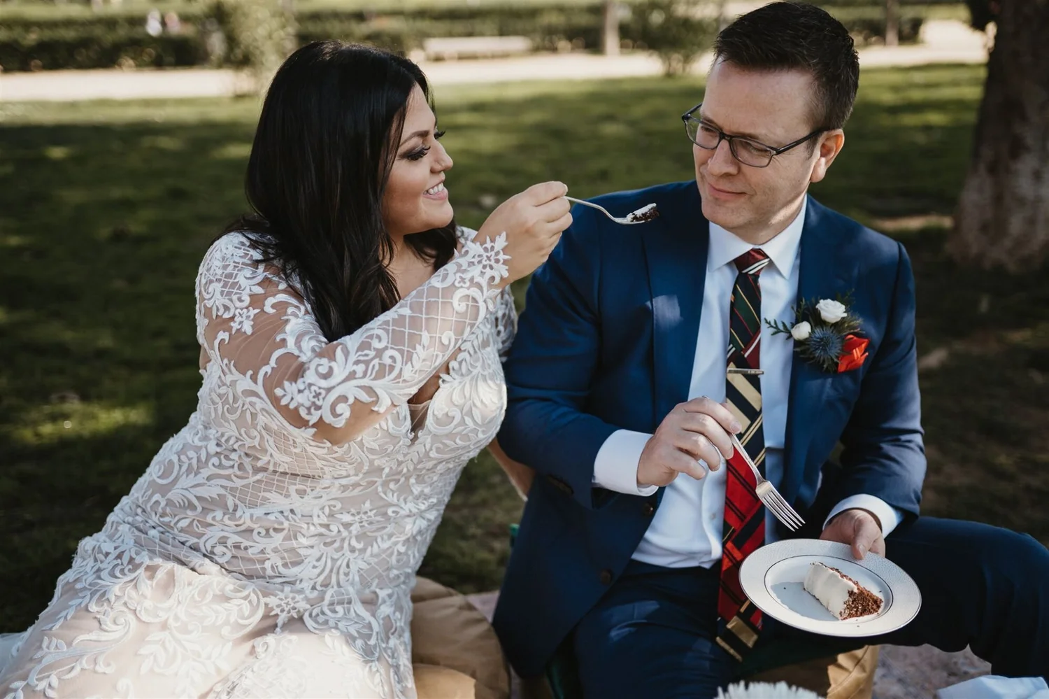 A woman in a wedding dress feeding a man in a suit with a spoon at an outdoor event
