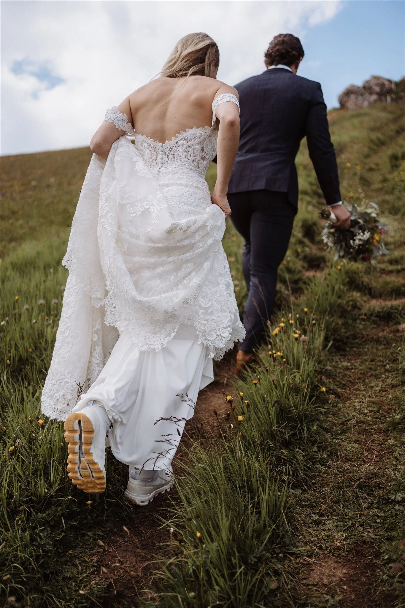 A bride in a white lace wedding dress and a groom in a dark suit walking up a grassy hill holding hands, with the groom holding a bouquet of flowers.