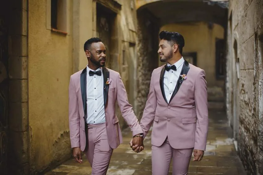 Two men wearing pink suits with black lapels, white shirts, and black bow ties, holding hands and walking through a narrow, rustic alleyway.