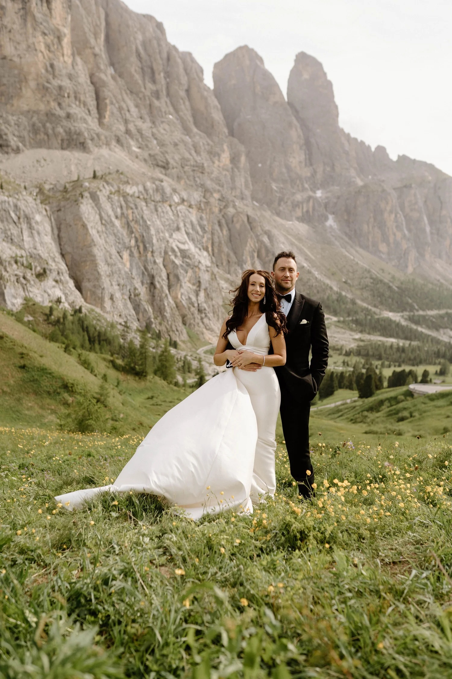 A bride and groom standing in a grassy field with yellow flowers, mountains in the background, the bride in a white wedding dress and the groom in a black tuxedo.