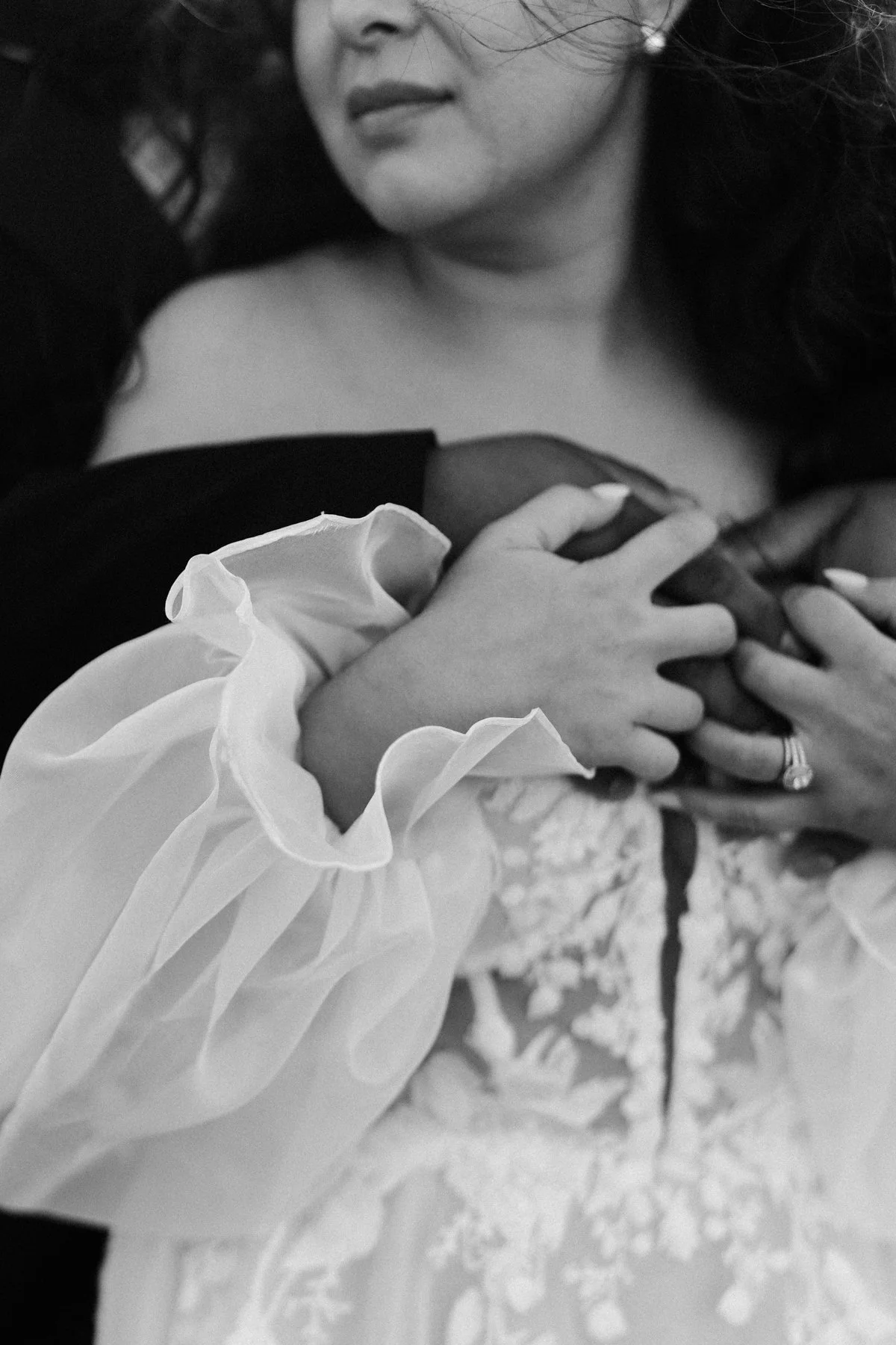 Close-up of a woman embracing a man, with her hand touching his chest. The woman is wearing a dress with a ruffled sleeve and a large ring on her finger. The image is in black and white.