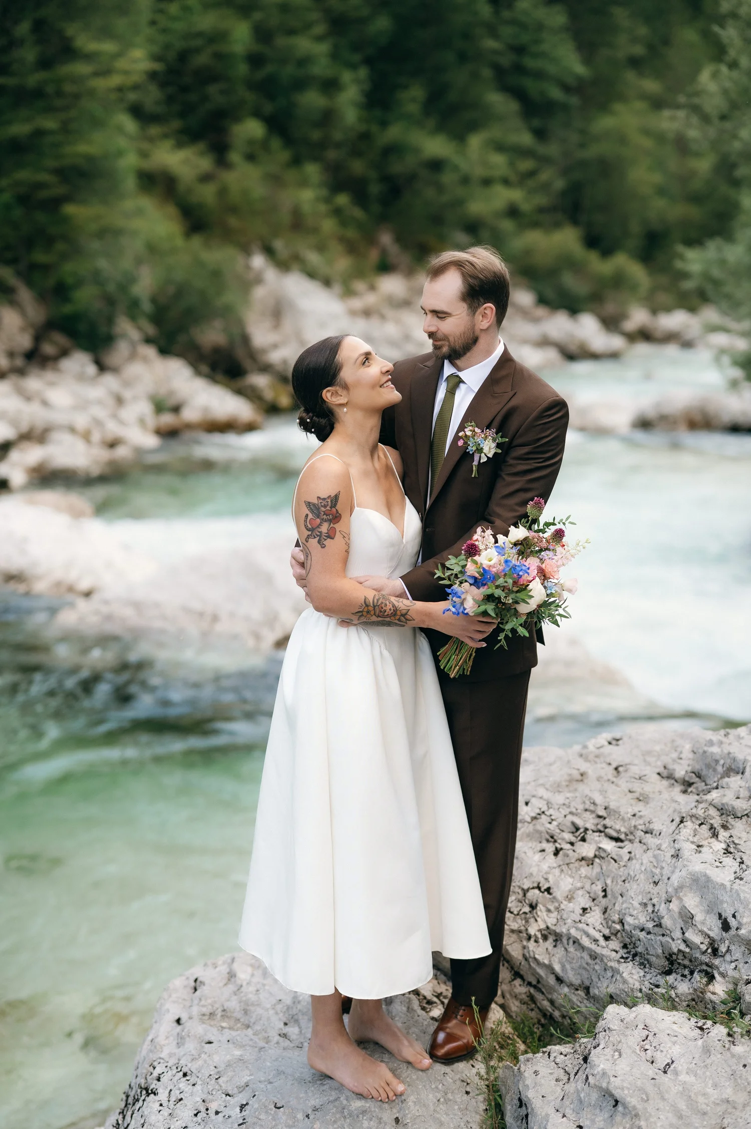 A couple is standing by a river with rocks and greenery in the background, embracing and smiling at each other. The woman is holding a bouquet of flowers, and she has tattoos on her arms. The man is wearing a brown suit and tie, and they both appear happy.