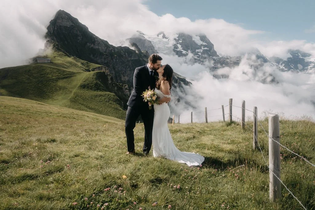 A bride and groom kiss on a grassy hillside with a mountainous background, clouds, and fence posts.
