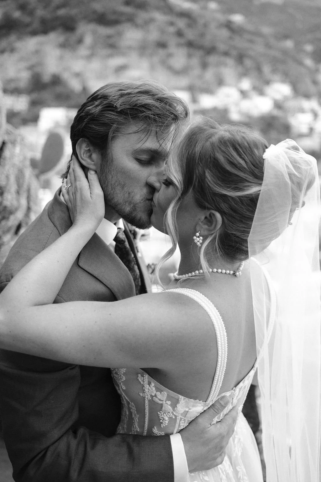 A black and white photo of a newlywed couple kissing outdoors, with a blurred hilly landscape in the background. The groom has short, wavy hair and a beard, dressed in a suit. The bride wears a wedding dress with lace details, a pearl necklace, earri
