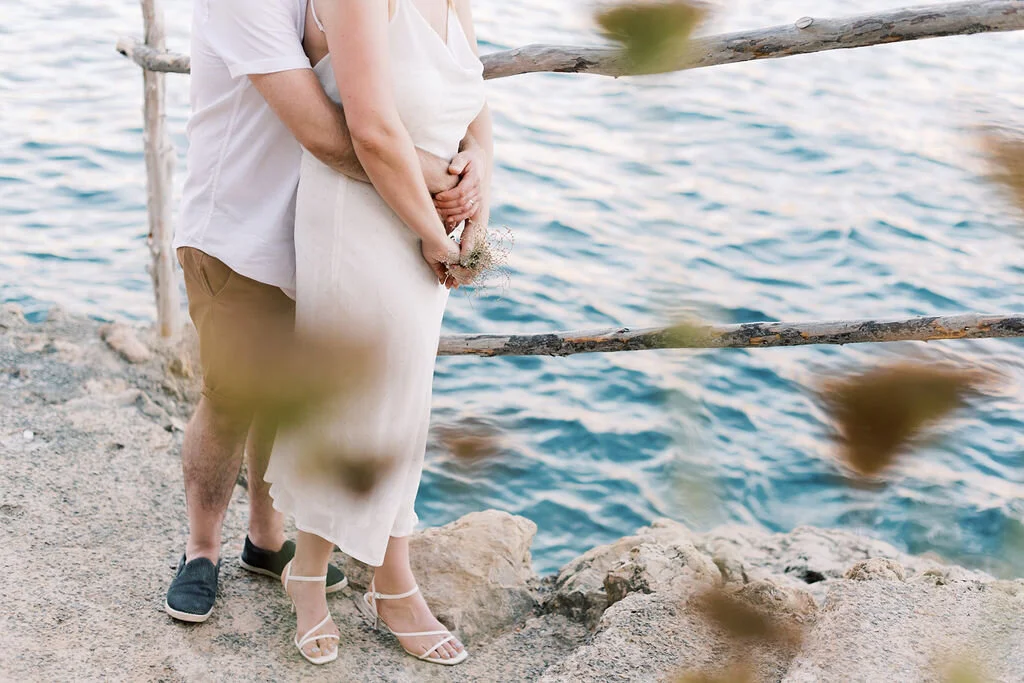 A couple standing near a body of water, with the man hugging the woman from behind. The woman is holding a small bouquet of flowers, and they are on a rocky shoreline. The scene is serene and romantic.