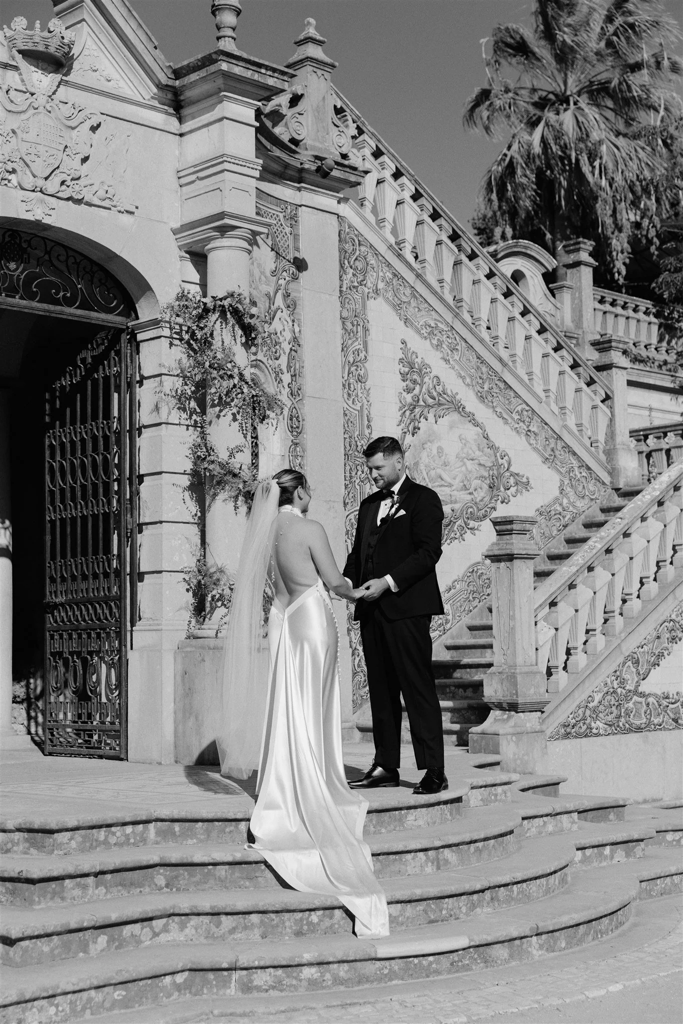 A wedding couple stands on the steps of a grand, ornate building with detailed carvings and elements of classical architecture. The bride wears a long, elegant white gown and veil, while the groom is dressed in a black tuxedo with a bow tie. They are