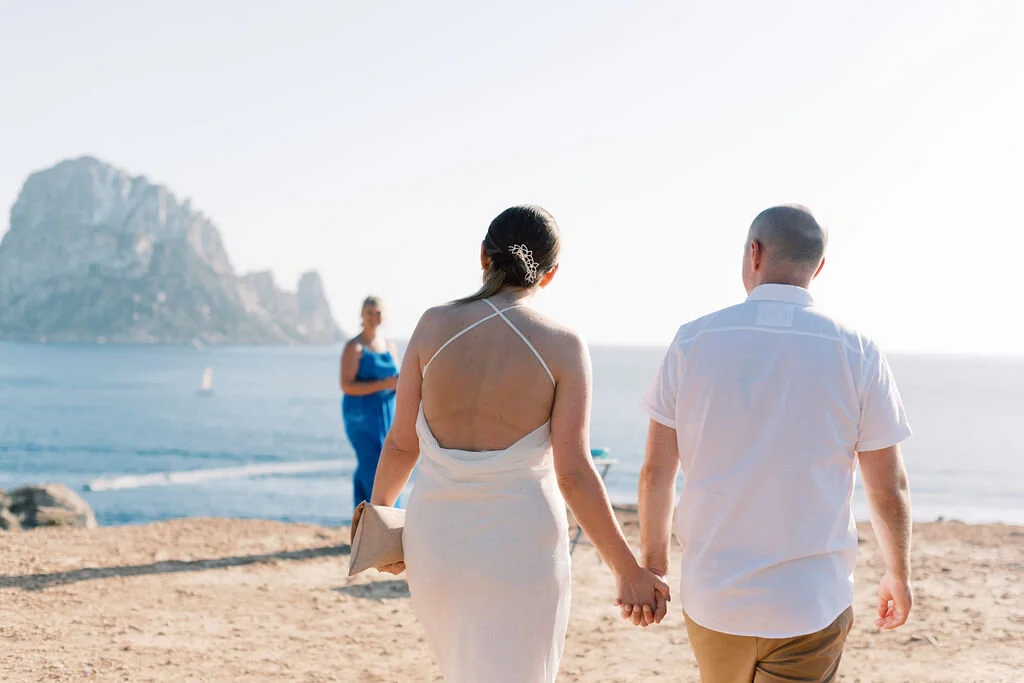 A couple holding hands walking on a beach with an ocean and a mountain in the background, while a woman in blue looks on.