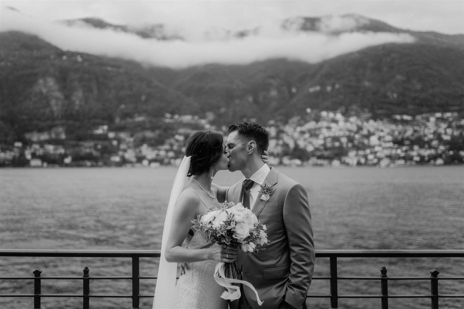 A black and white photo of a bride and groom sharing a kiss outdoors, with a body of water and mountains in the background.