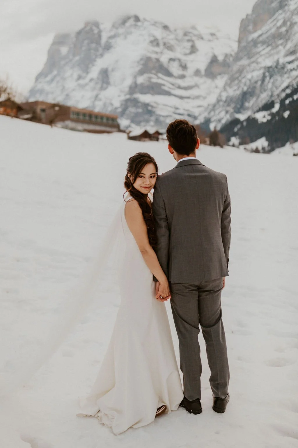 A bride and groom standing hand-in-hand in a snowy landscape with mountains in the background, with the bride smiling and looking at the camera.