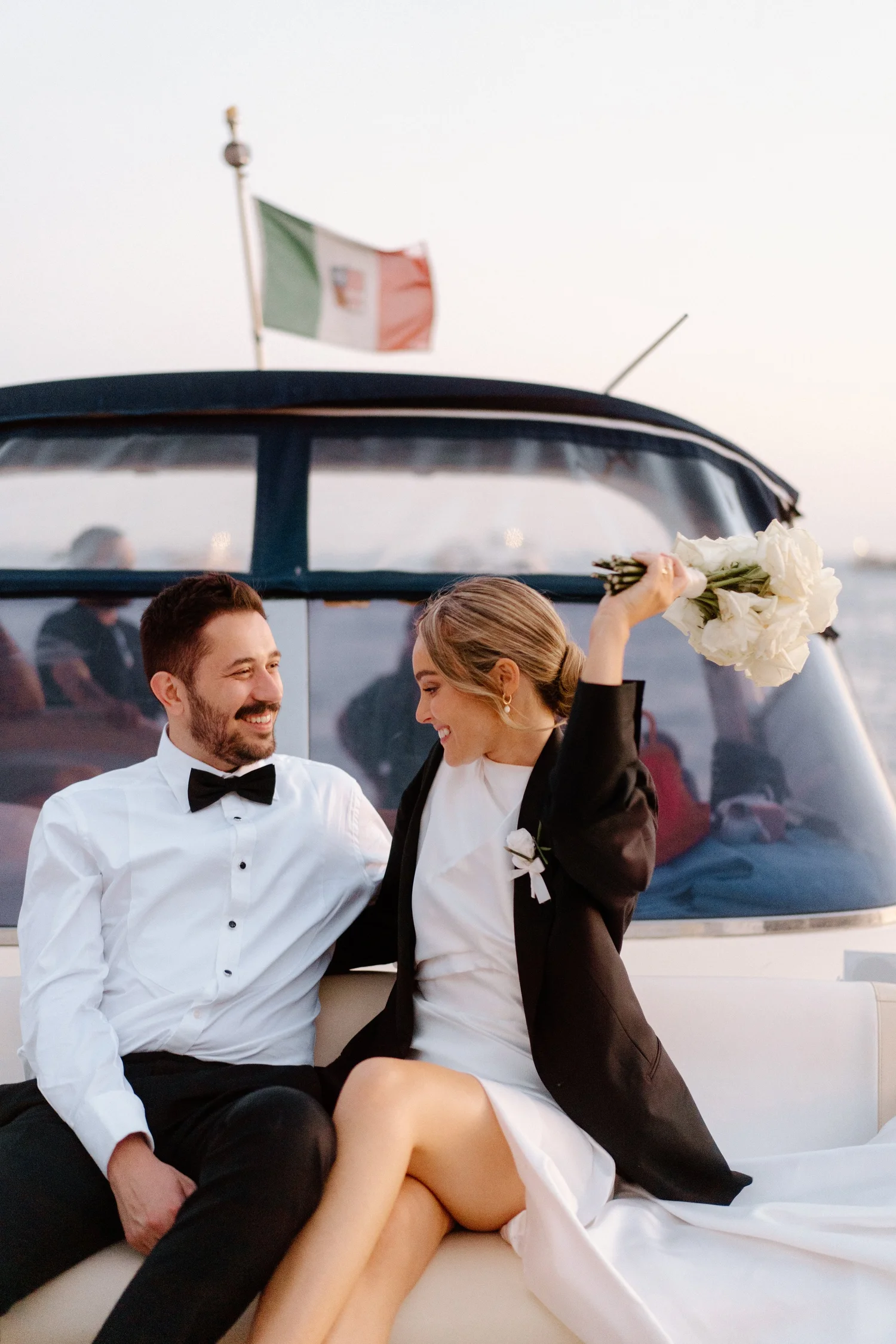 A newlywed couple sitting together on a yacht, smiling. The woman is holding a bouquet of white flowers, and the man is wearing a tuxedo with a bow tie. An Italian flag is flying on the yacht in the background.