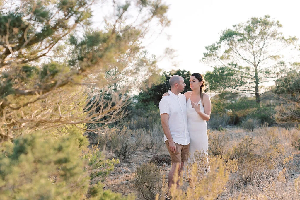 A couple standing close together outdoors in a natural setting with trees and shrubs, during sunset or late afternoon.