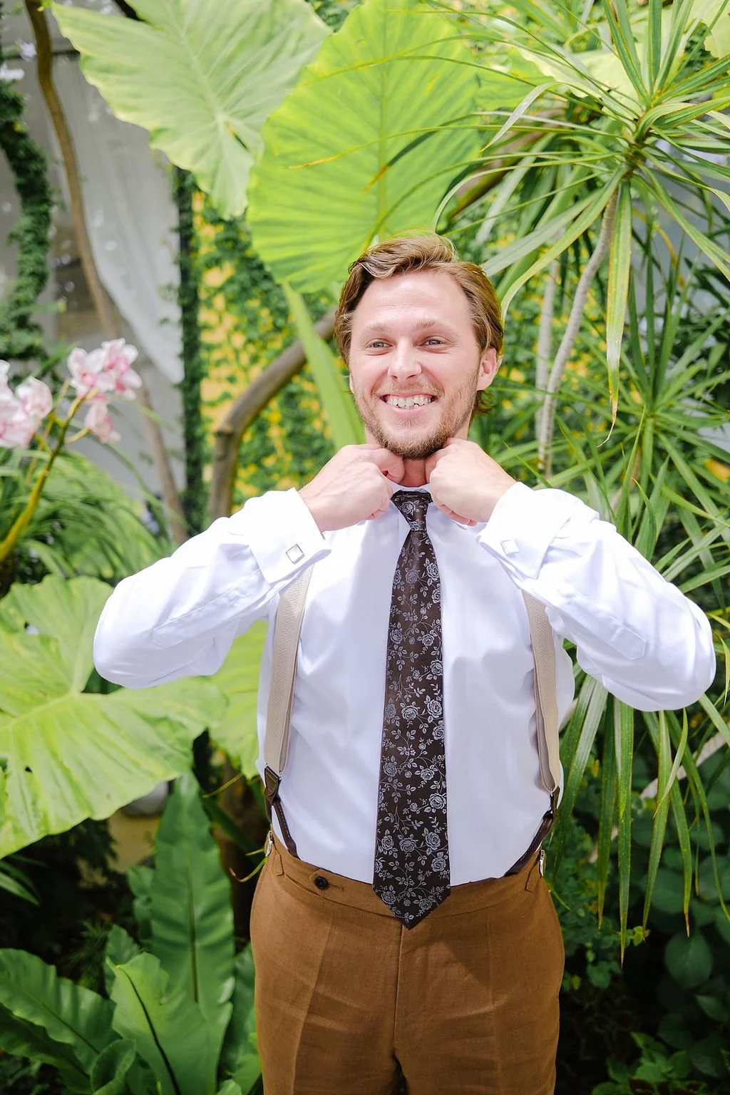 Man in white shirt and patterned tie adjusting suspenders in a lush, green tropical garden.