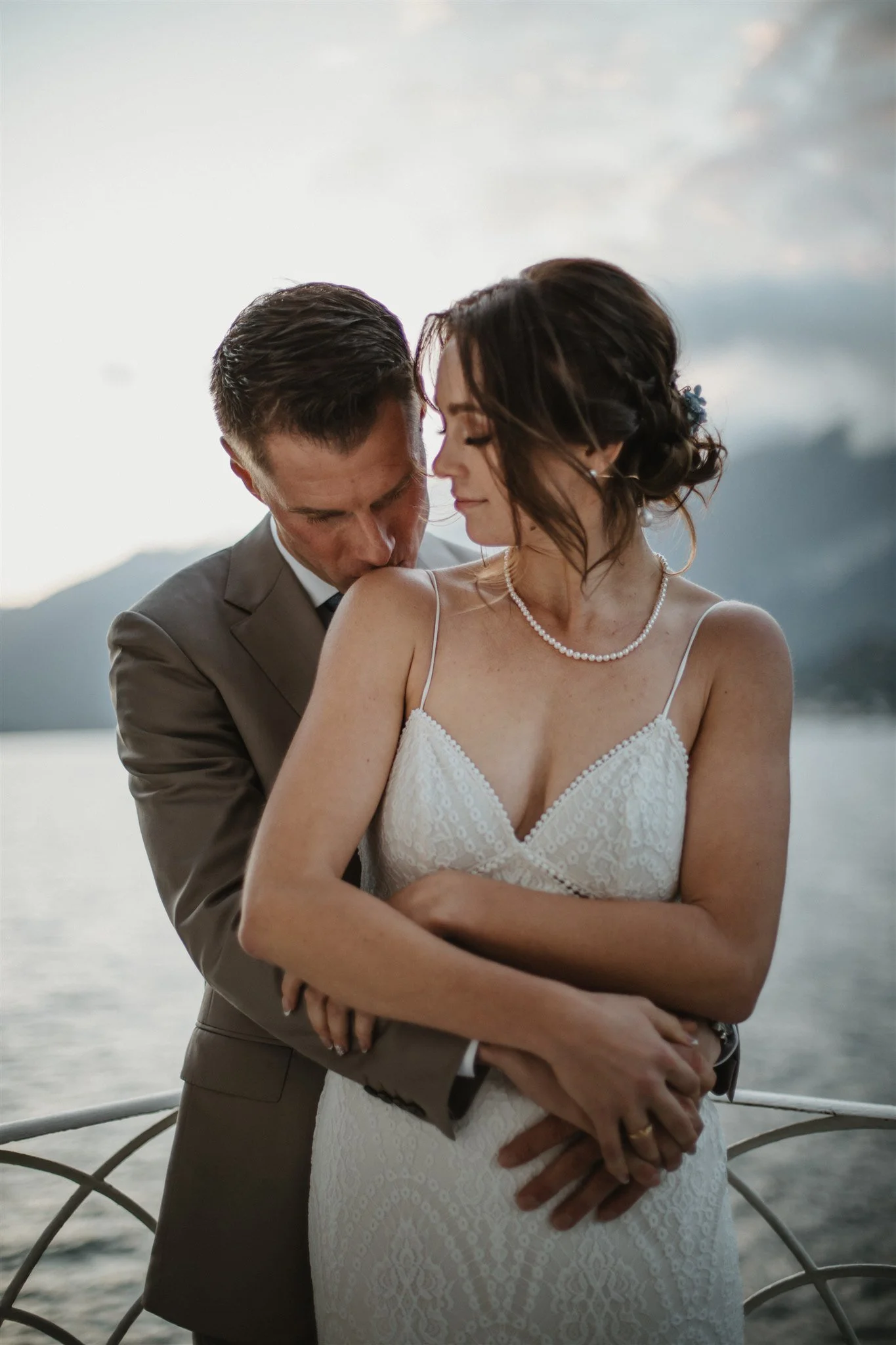 A couple dressed in wedding attire standing closely on a boat or dock with mountains and water in the background at sunset or dusk.
