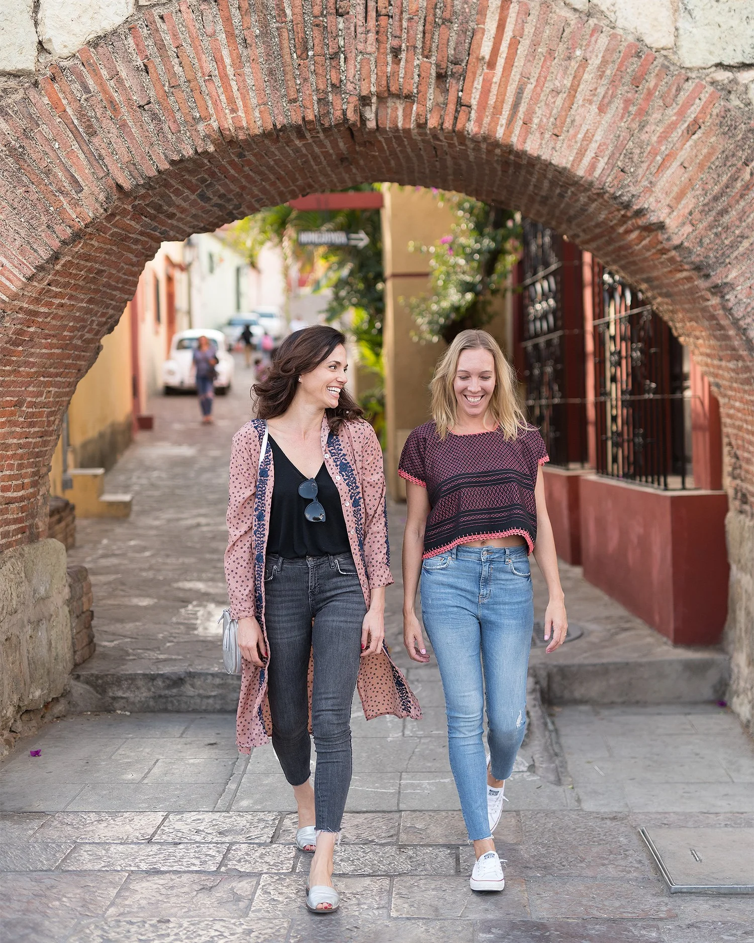 Two women walking and talking under a brick archway on a cobblestone street in a colorful neighborhood, smiling and enjoying each other's company.