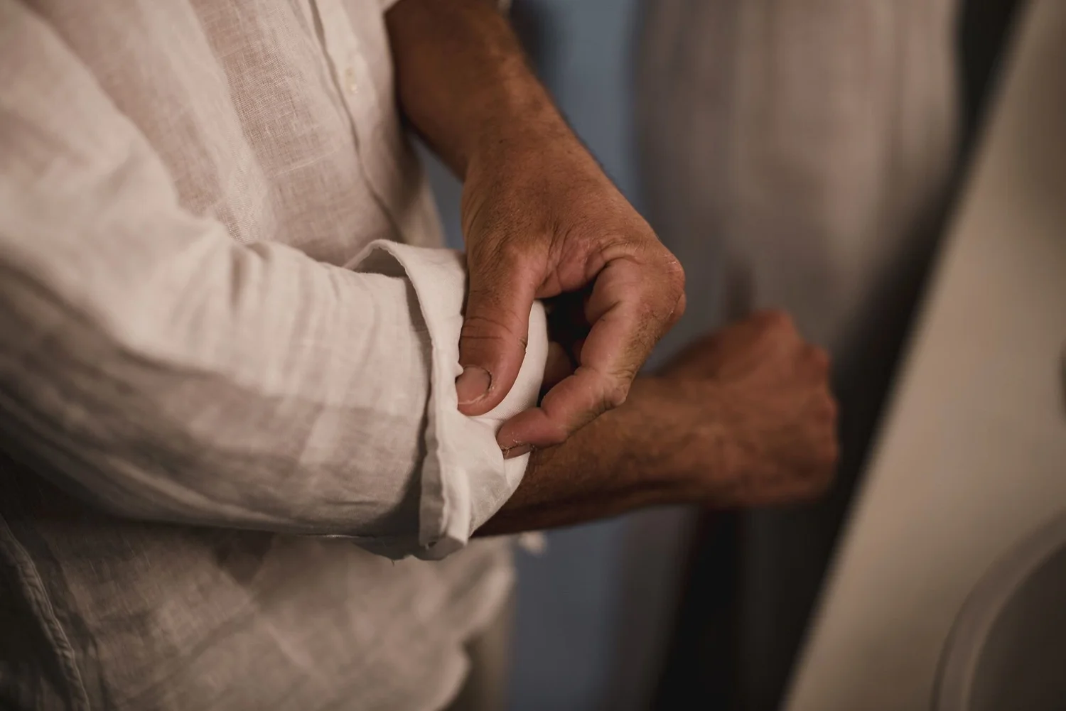 Close-up of a person's arm with tanned skin, wearing a beige, long-sleeved shirt. The person is resting their hand on their lap, fingers slightly curled.
