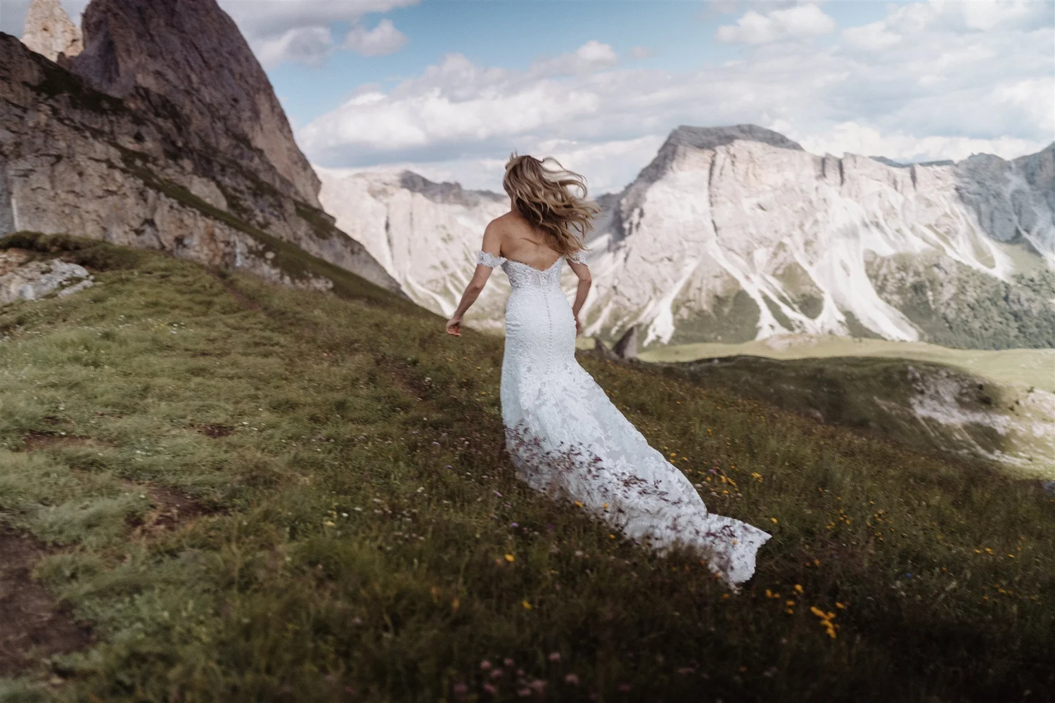 A woman in a white lace wedding dress walking through a grassy mountain landscape with rugged, snow-capped mountains in the background under a partly cloudy sky.