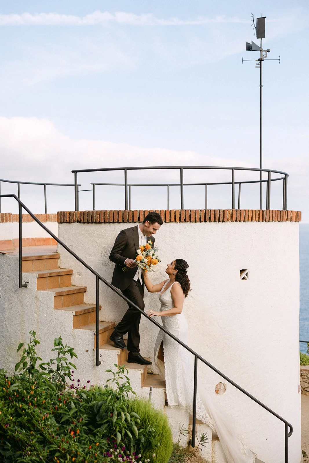 A bride and groom on a white staircase outside, with the groom giving the bride a bouquet of flowers, under a cloudy sky.
