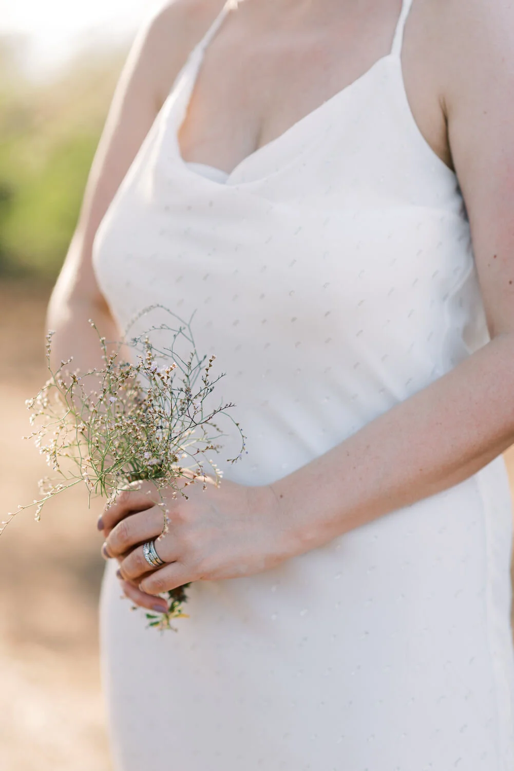 A woman in a white dress holding a small bouquet of wildflowers, with her hand showing a wedding ring.