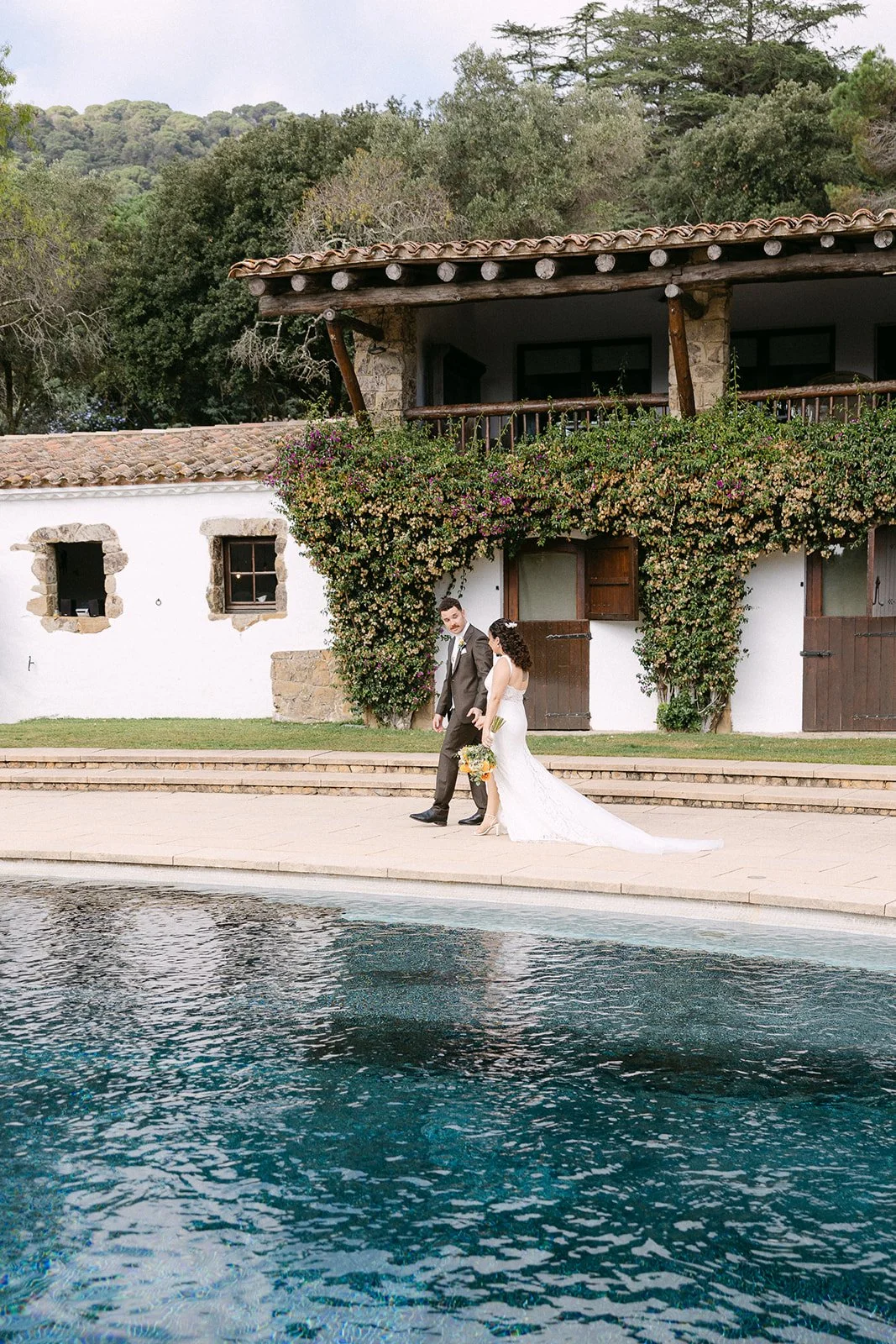 Wedding couple walking by a pool outside a rustic building with climbing plants and a balcony