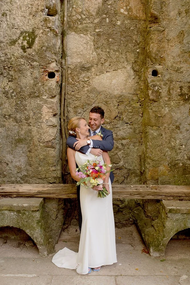 A bride and groom embracing, with the groom in a dark suit and the bride in a white dress holding a bouquet of colorful flowers, standing against an old stone wall.
