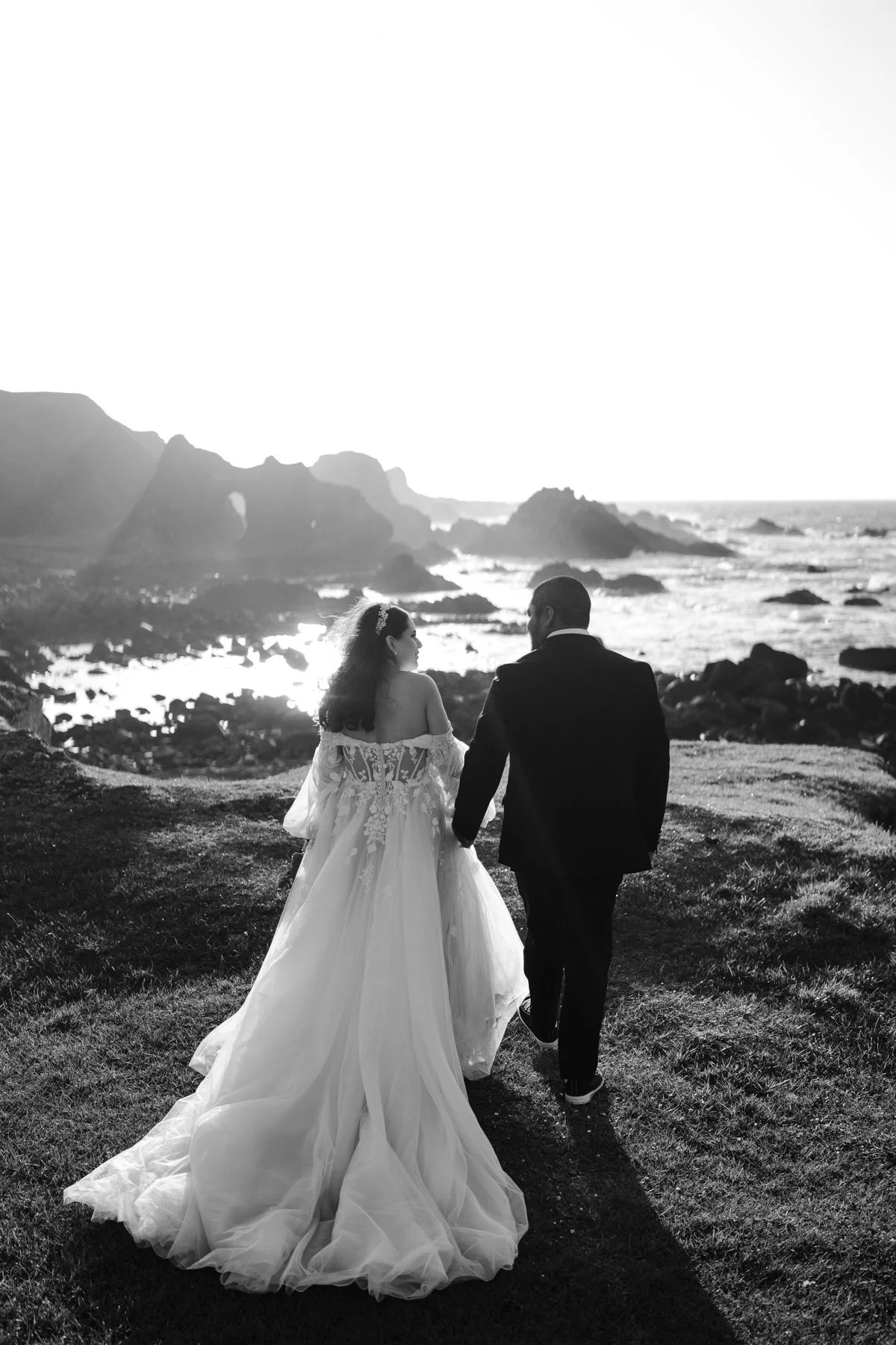 Black and white photo of a bride and groom walking hand in hand on a grassy hill near the ocean, with rocky cliffs in the background, during sunset.