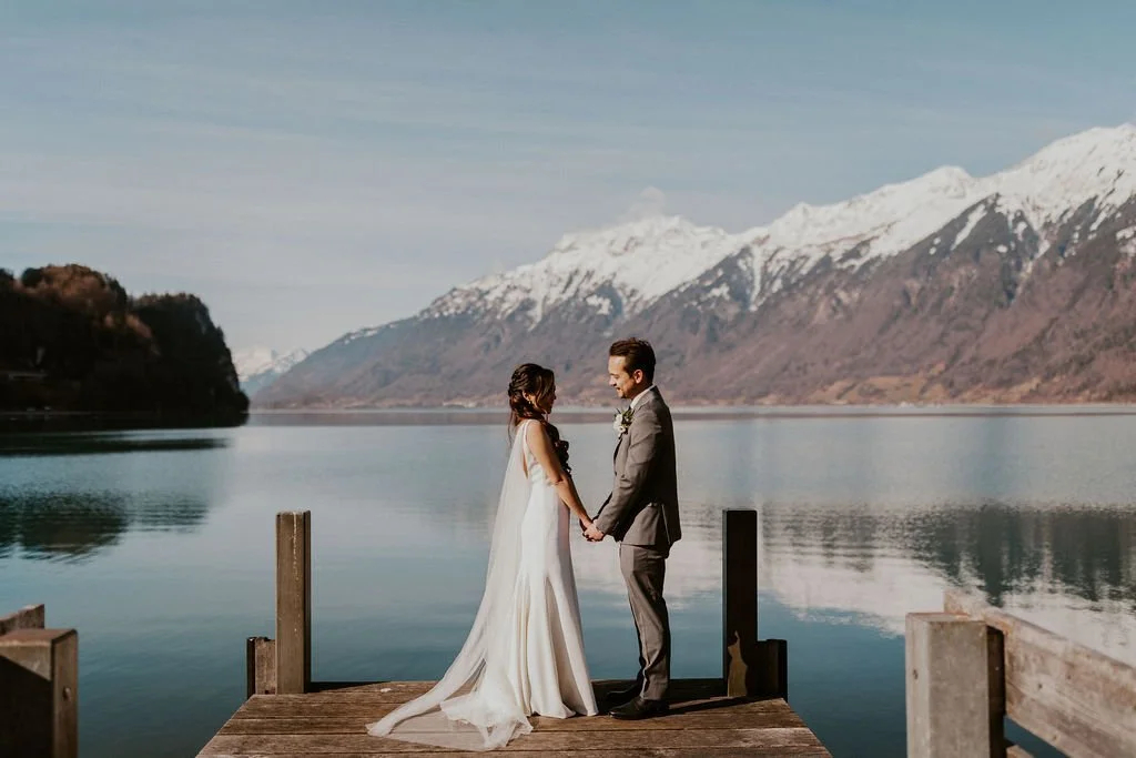 A bride and groom holding hands on a wooden dock by a calm lake with snow-capped mountains in the background.