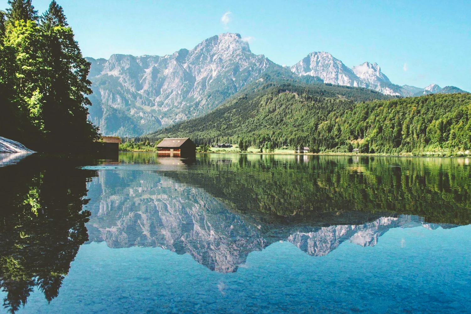 A scenic view of a mountain landscape with lush green forests and a lake in the foreground, reflecting the mountains and sky.