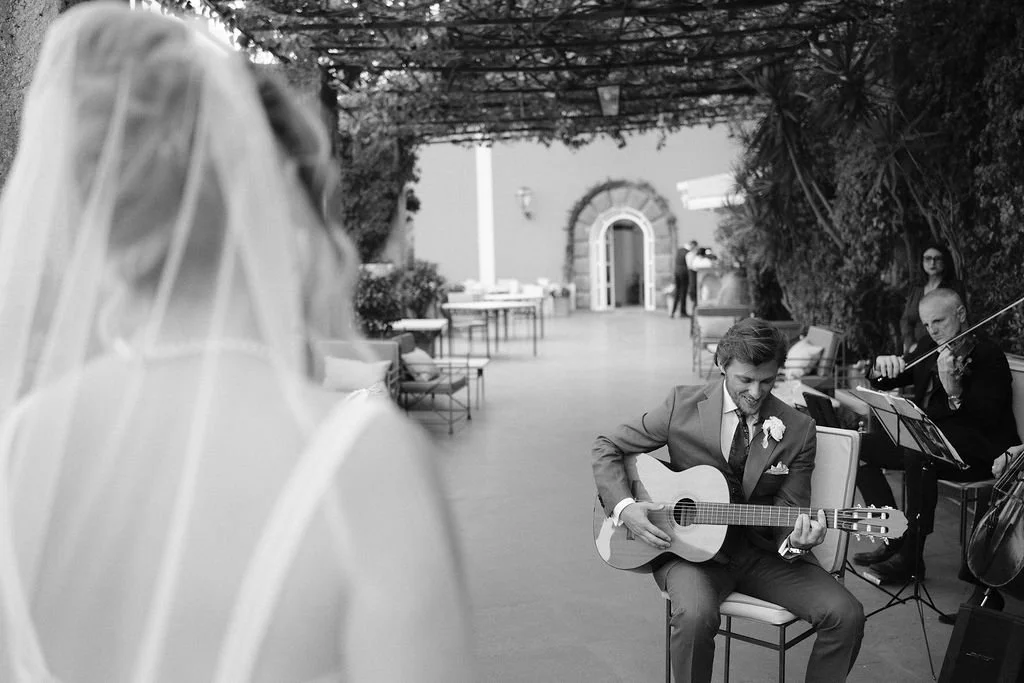 A black and white photo of a wedding ceremony. A bride with a veil is standing in the foreground, facing a groom who is playing an acoustic guitar. A musician is playing a violin in the background. The setting is an outdoor patio with chairs and tabl
