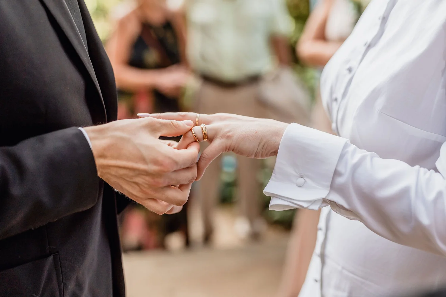 A couple exchanging wedding rings during a ceremony, with the focus on their hands and rings, and blurred guests in the background.