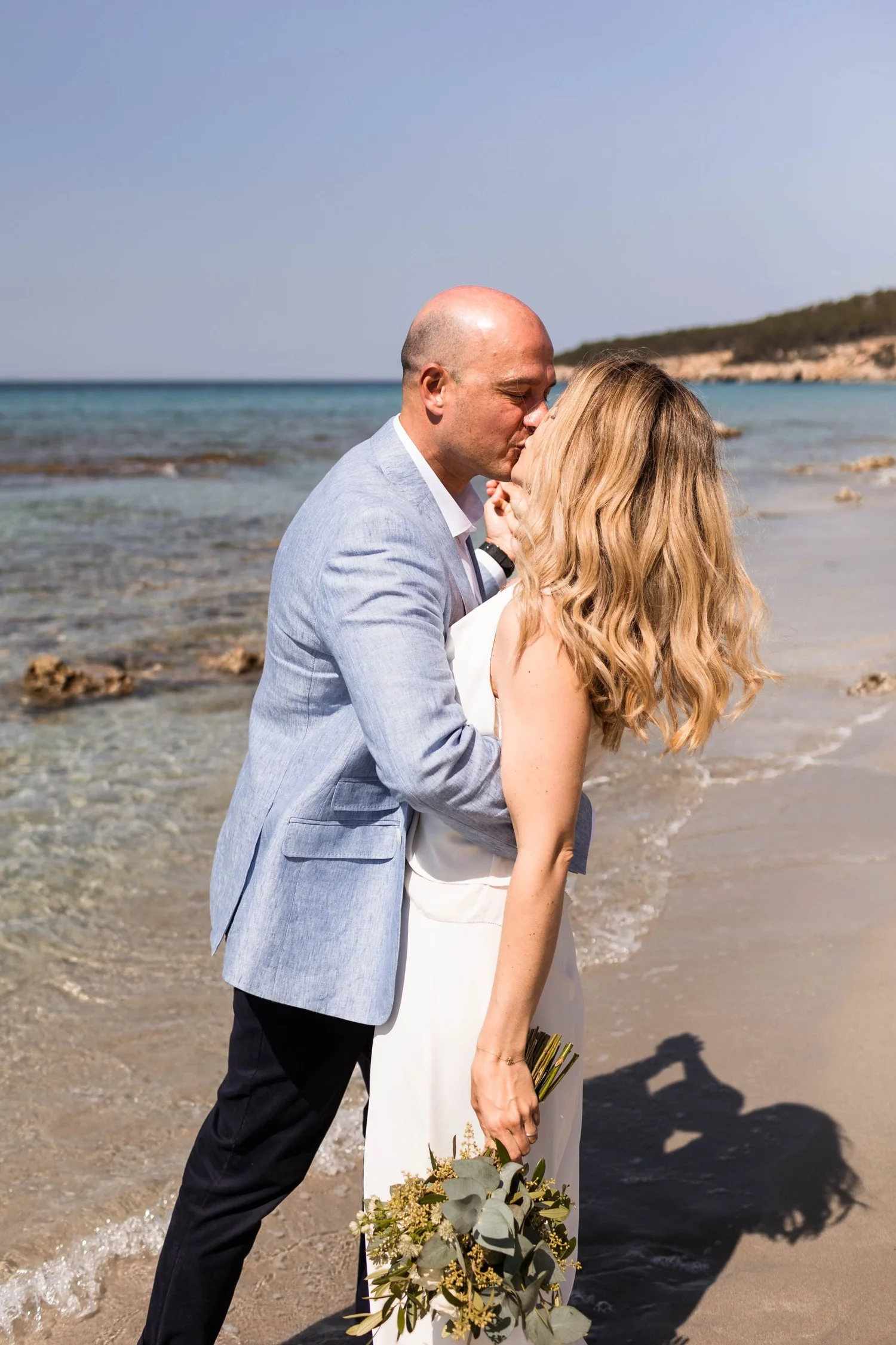 A couple dressed in wedding attire sharing a kiss on the beach near the water with a distant shoreline and clear sky.