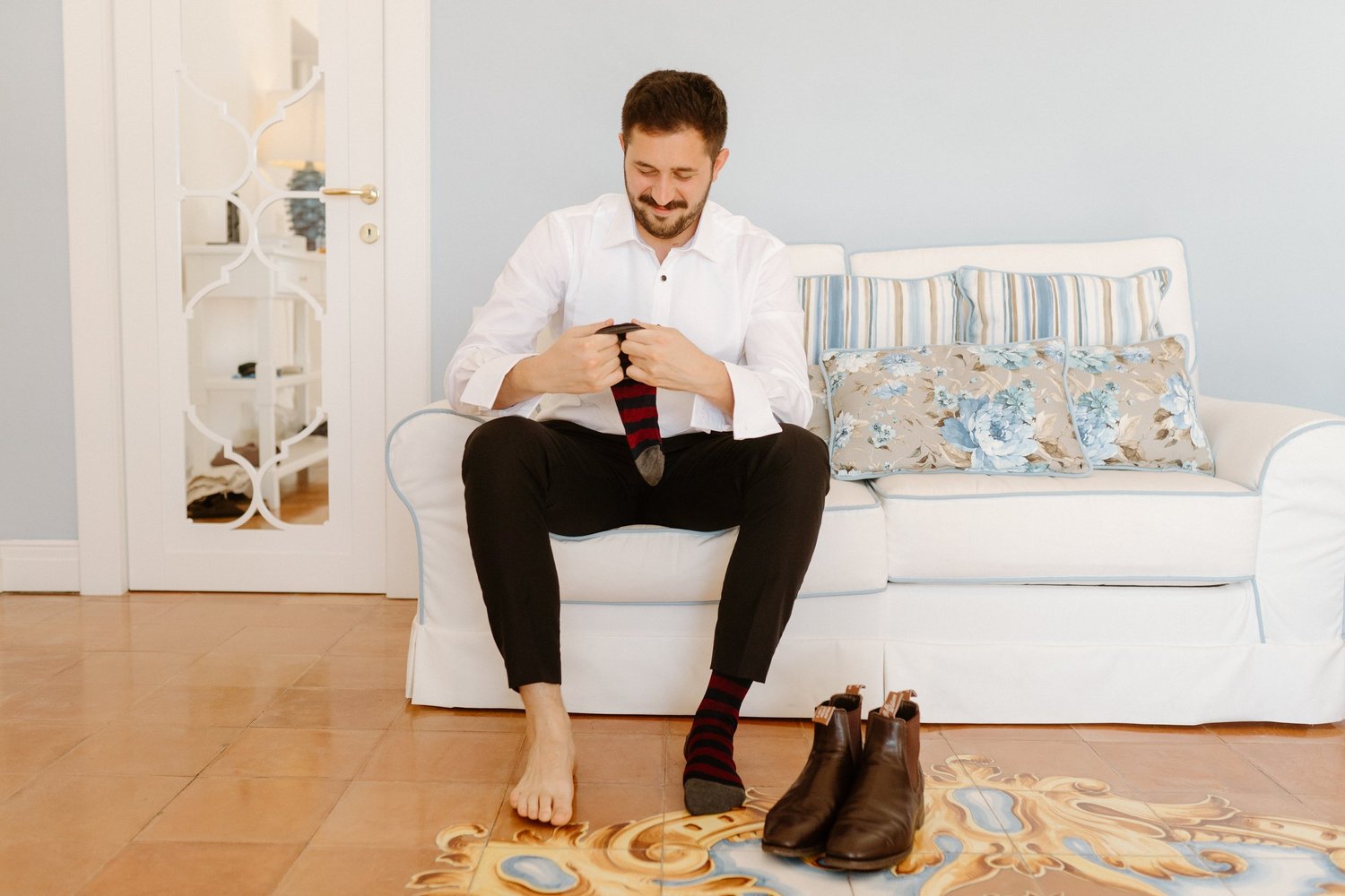 A man sitting on a white sofa, smiling and looking at his phone. He is in a room with a decorative mirror, some pillows with floral and striped patterns, and brown shoes on the floor. He is in the process of putting on or taking off socks, with one s