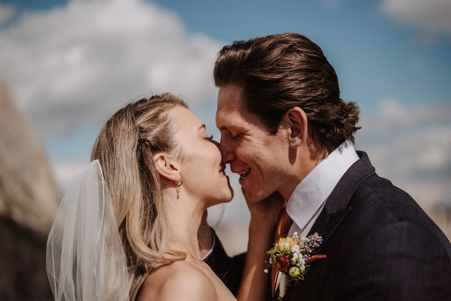 A bride and groom share a close moment, about to kiss, outdoors under a sky with clouds.