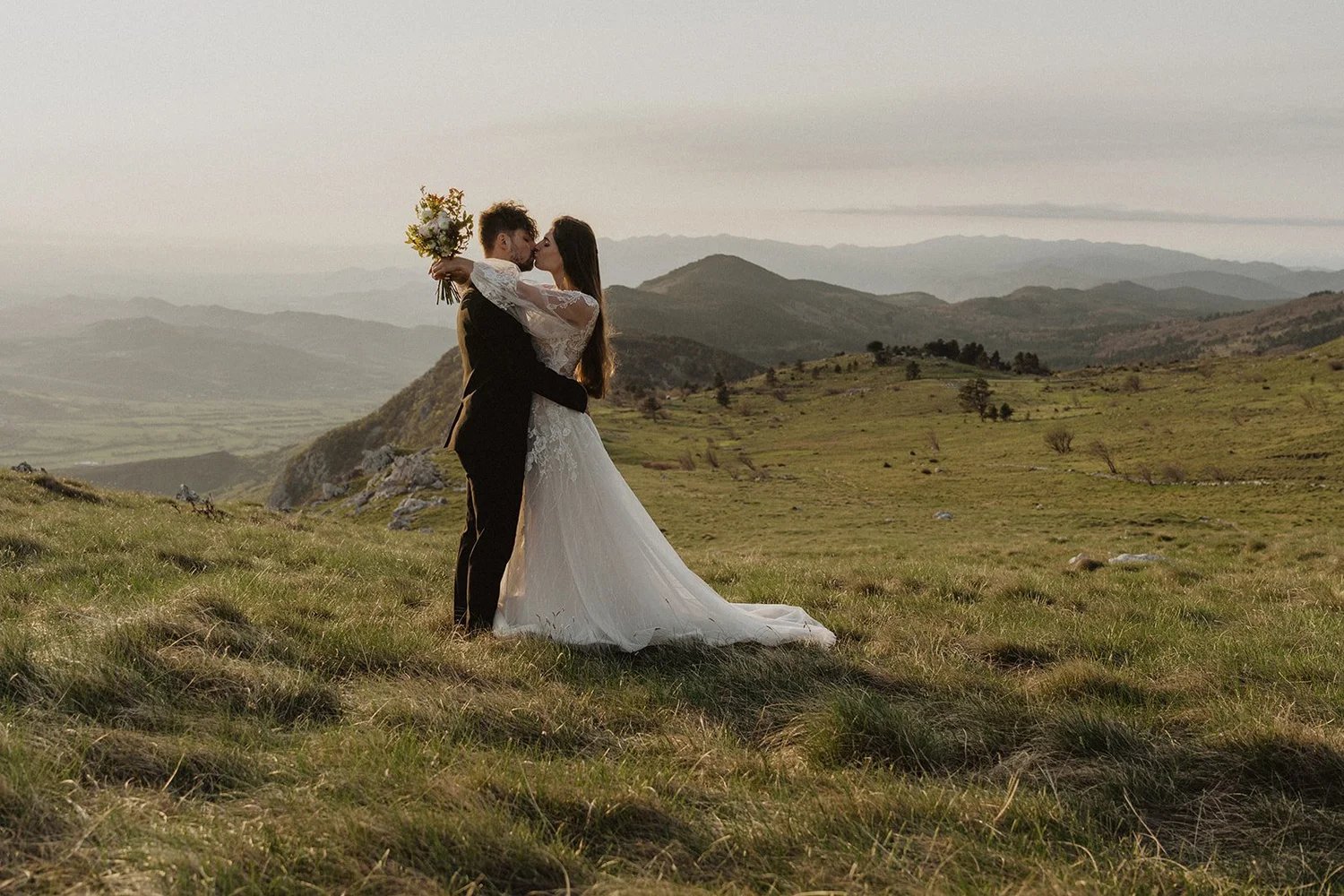 A couple in wedding attire kissing in a grassy field with rolling hills and mountains in the background during sunset.