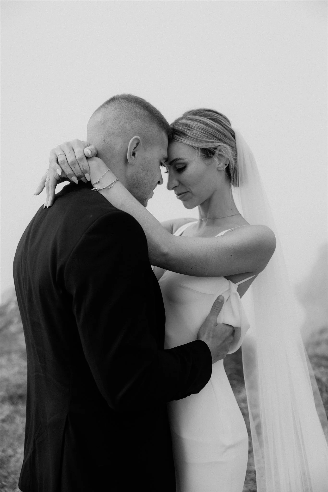 A black and white photo of a couple embracing, standing close with foreheads touching, eyes closed, and gentle smiles. The man is wearing a dark suit, and the woman is wearing a light dress with a veil. Their hands are gently placed on each other's bodies, showing intimacy and love.